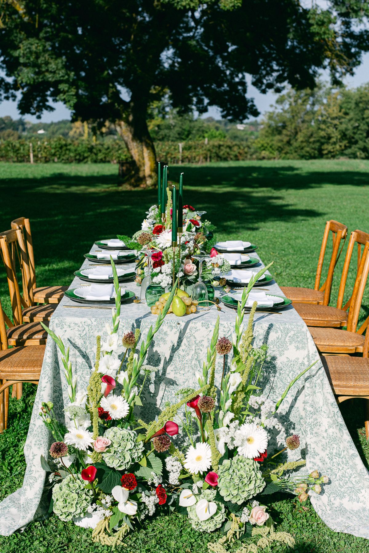 A long table with patterned linen, wooden chairs and large red, white and green floral arrangements in bright sunlight.