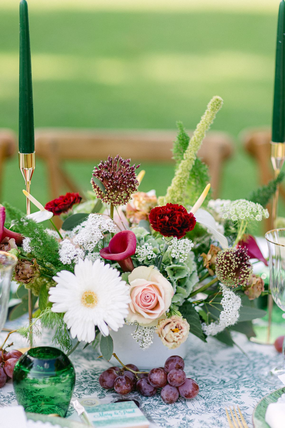 A table centrepiece of white, pink and red flowers with tall green candles, a green glass vase and a bunch of dark grapes.