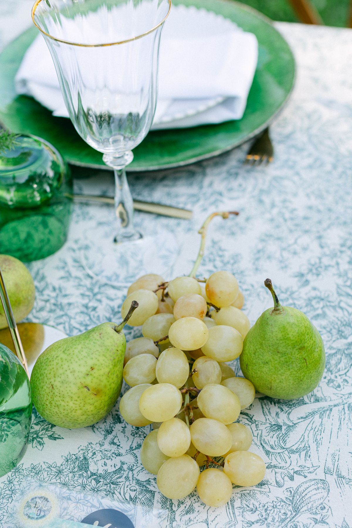 Pears and grapes on a green toile tablecloth beside a gold-rimmed glass