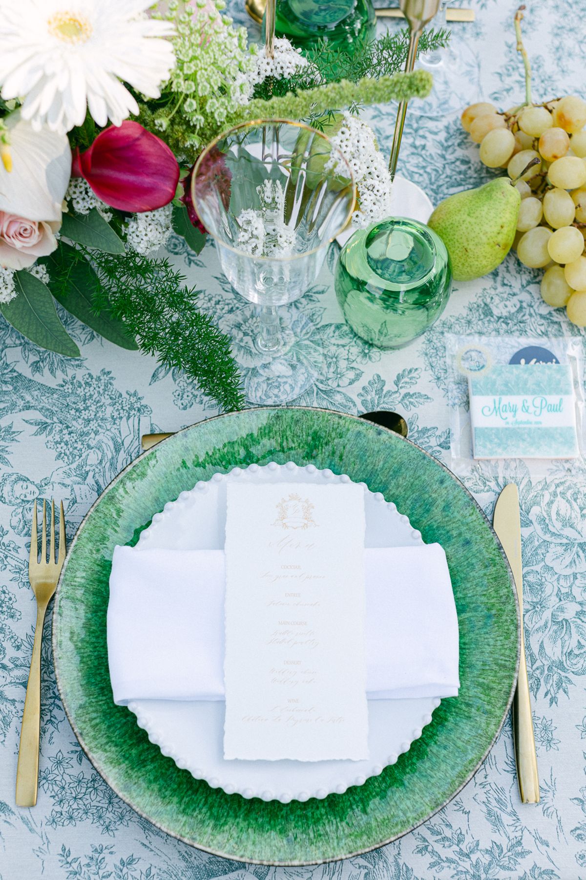 A table setting with a green scalloped plate, folded white menu, gold cutlery, grapes, pears and colourful flowers on patterned linen.
