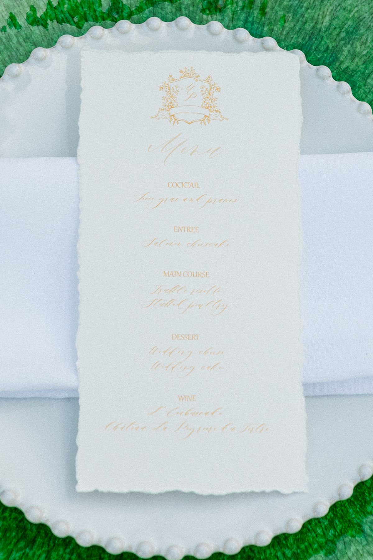 A cream menu card with a gold crest and handwritten calligraphy rests on a folded white napkin and a beaded-rim plate, shot from above.