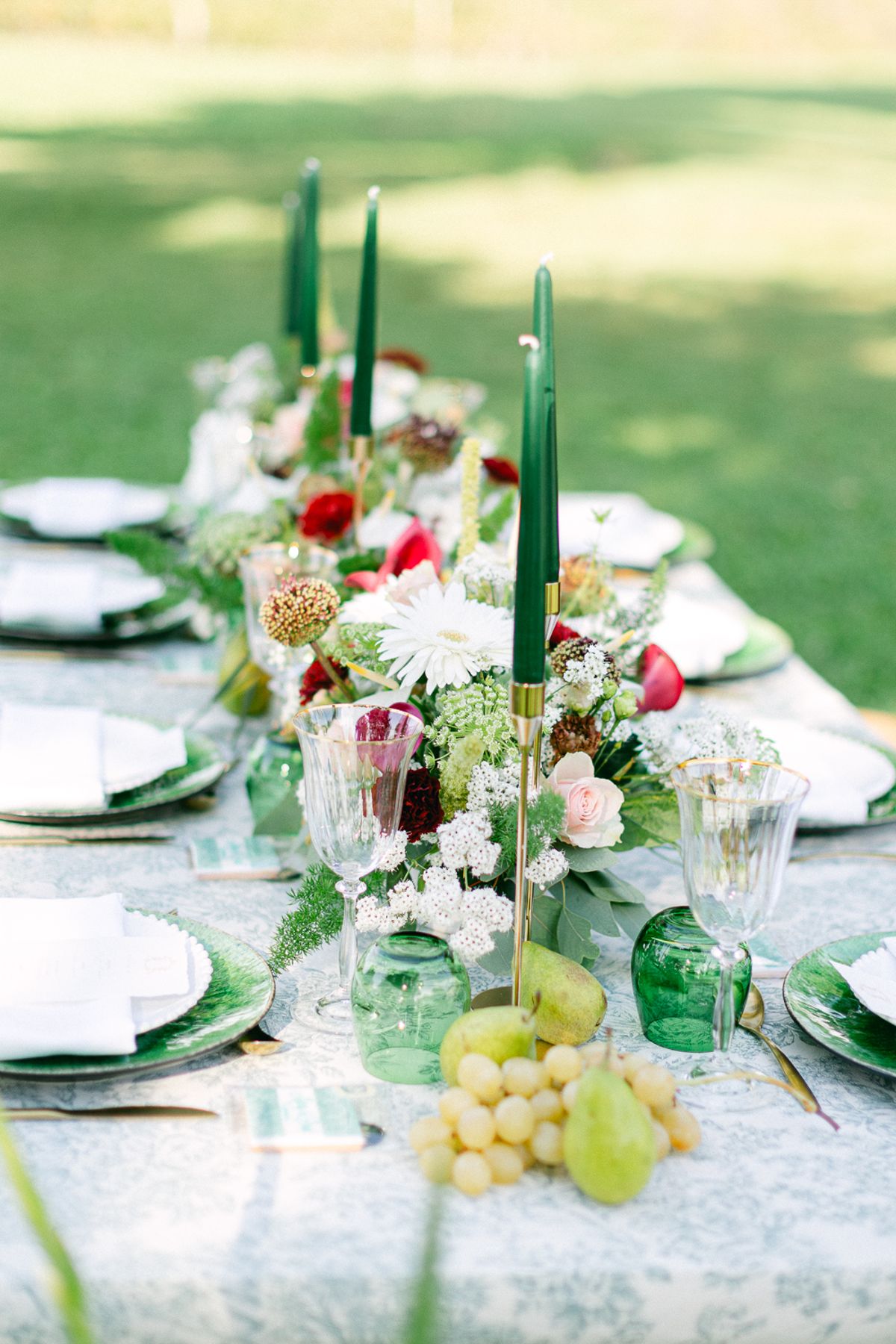 A table decorated with tall green candles, grapes, pears, green glassware and flowers, with plates in soft focus.