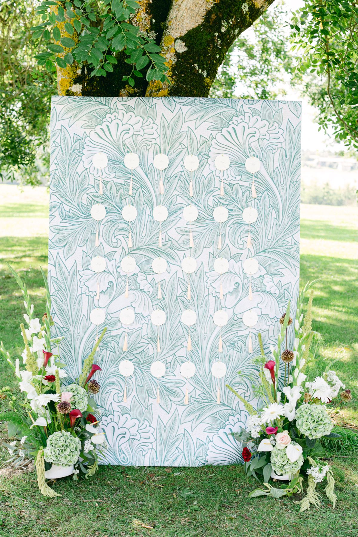 A tall pale green patterned seating board stands on a lawn beneath a tree, flanked by arrangements of white and red flowers.