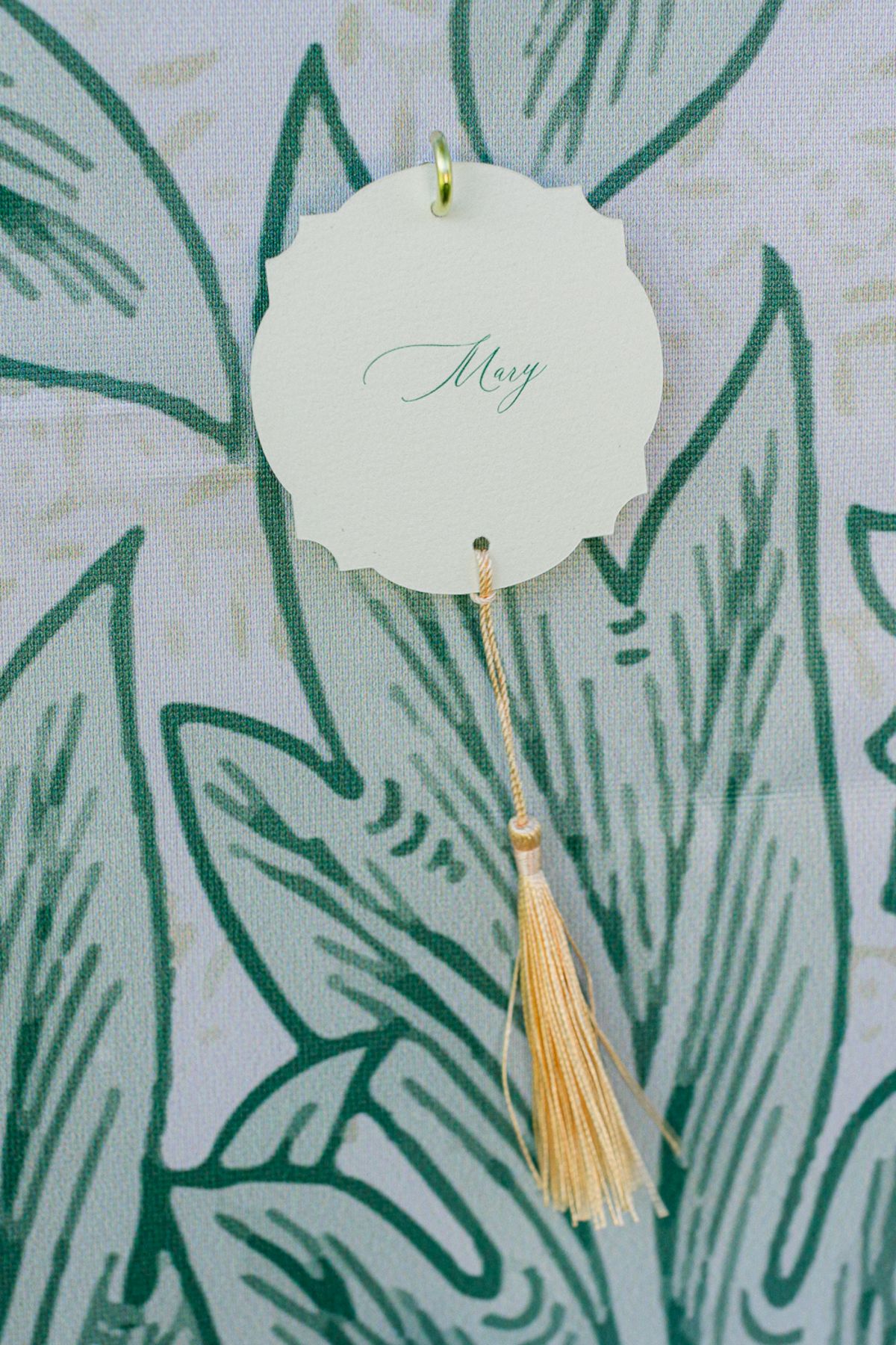 A round cream place card with green calligraphy and a gold tassel rests against a green leaf-patterned surface.