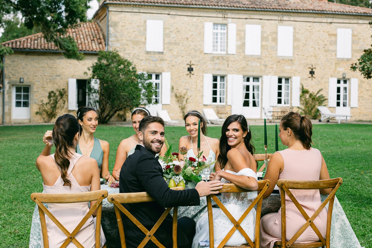 The groom and several women in pastel dresses sit at a wooden-chaired table on a lawn in front of a stone house.