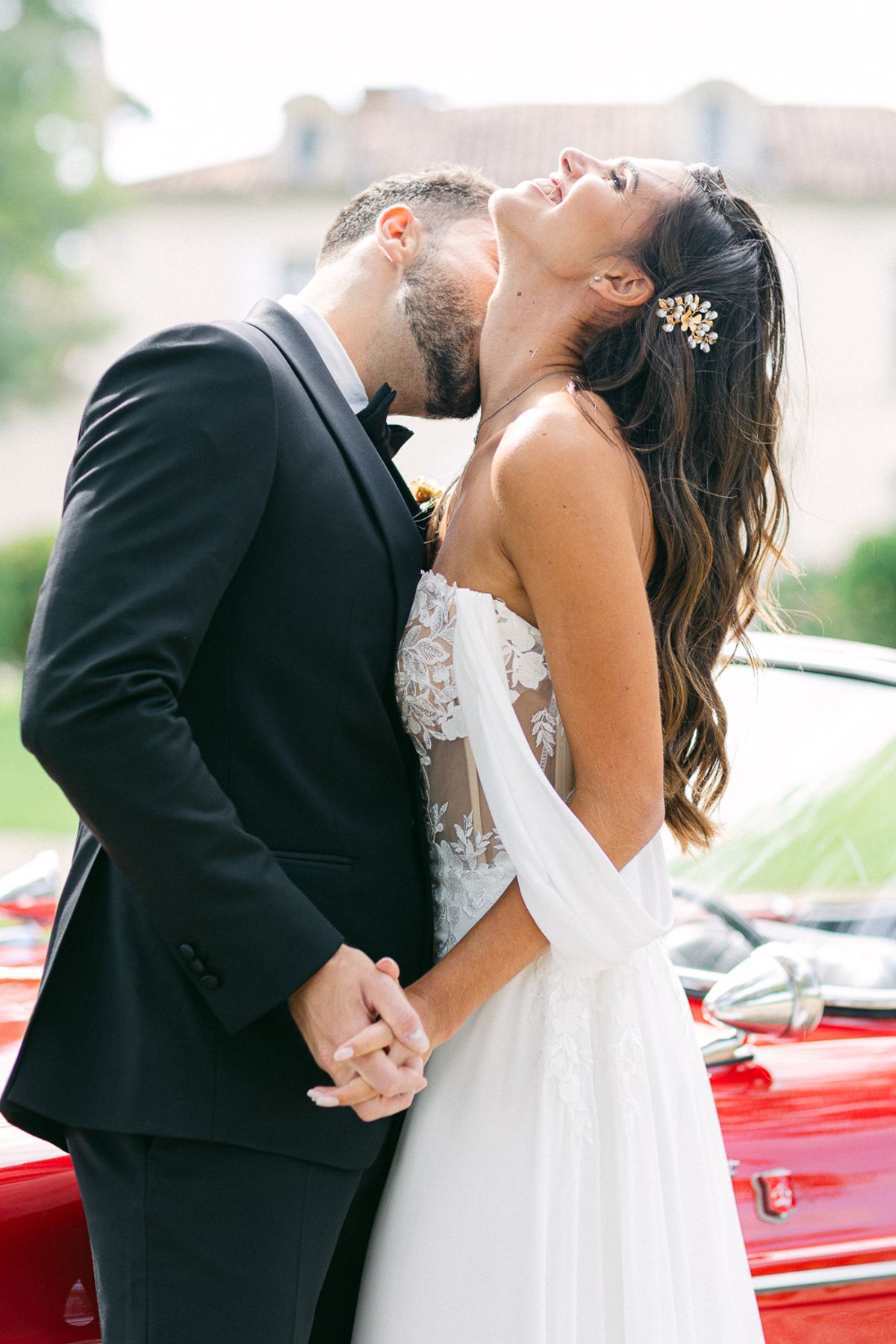 The groom in a black suit kisses the bride's neck as she tilts her head back laughing, their hands clasped beside a red car.