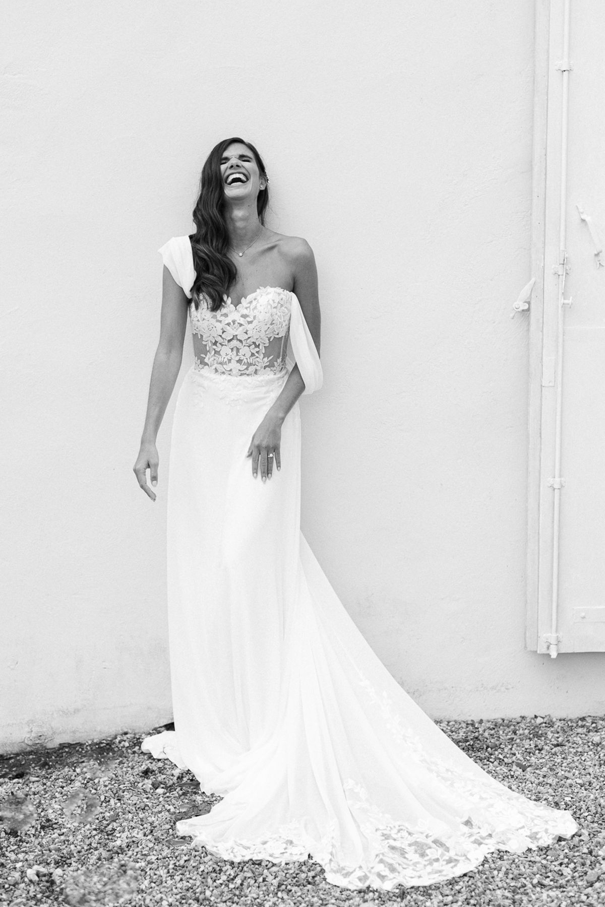 A black and white image of the bride laughing in a lace gown with a long train, standing against a plain wall.