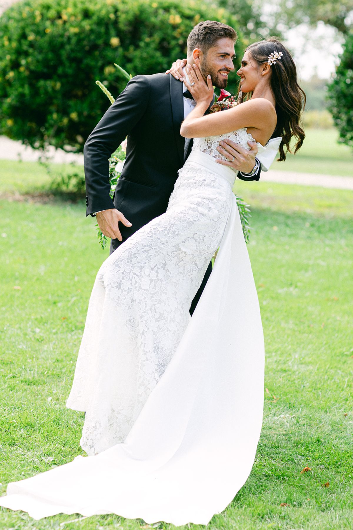 The groom in a black suit leans the bride back in an embrace on a green lawn, her lace dress and long train flowing.