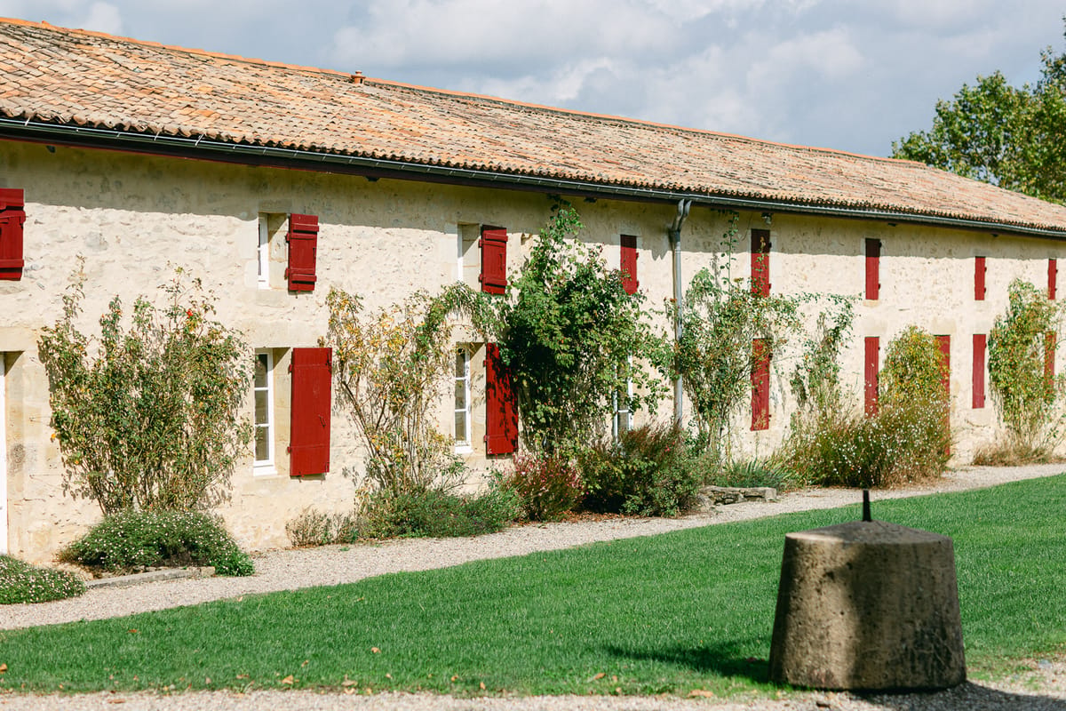 A long stone building with a tiled roof and red shutters, climbing plants and a green lawn in front.