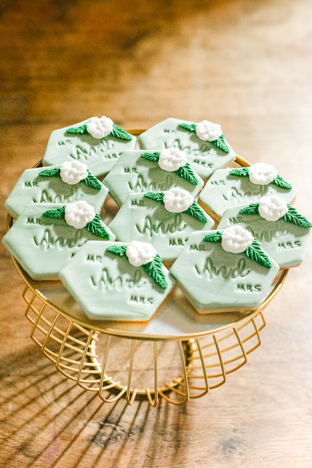 Hexagonal pale green biscuits decorated with white sugar flowers and leaves sit on a gold wire cake stand.