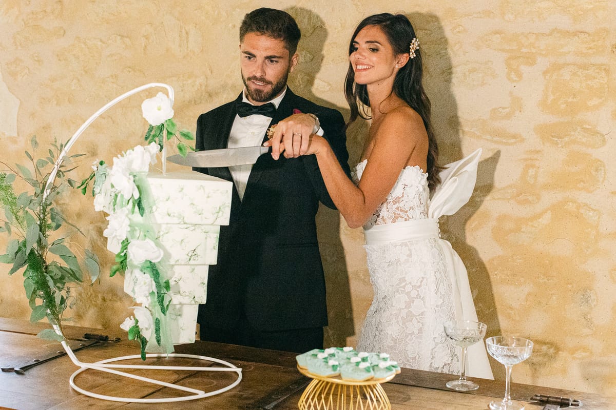 The groom in a black tuxedo and the bride in a lace gown cut a white tiered cake on a stand.