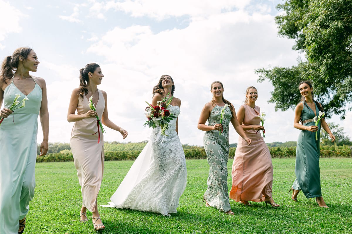 The bride and five bridesmaids in pastel dresses walk together across a green lawn beneath a blue sky with clouds.