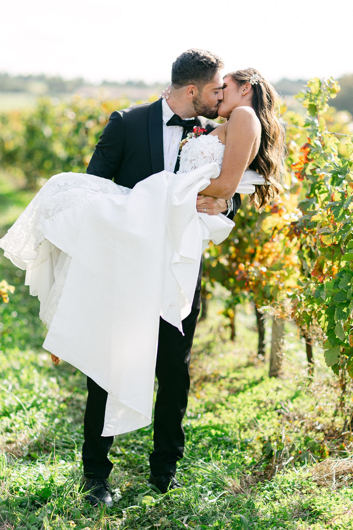 The groom in a black tuxedo lifts the bride in her white gown and kisses her in a vineyard.