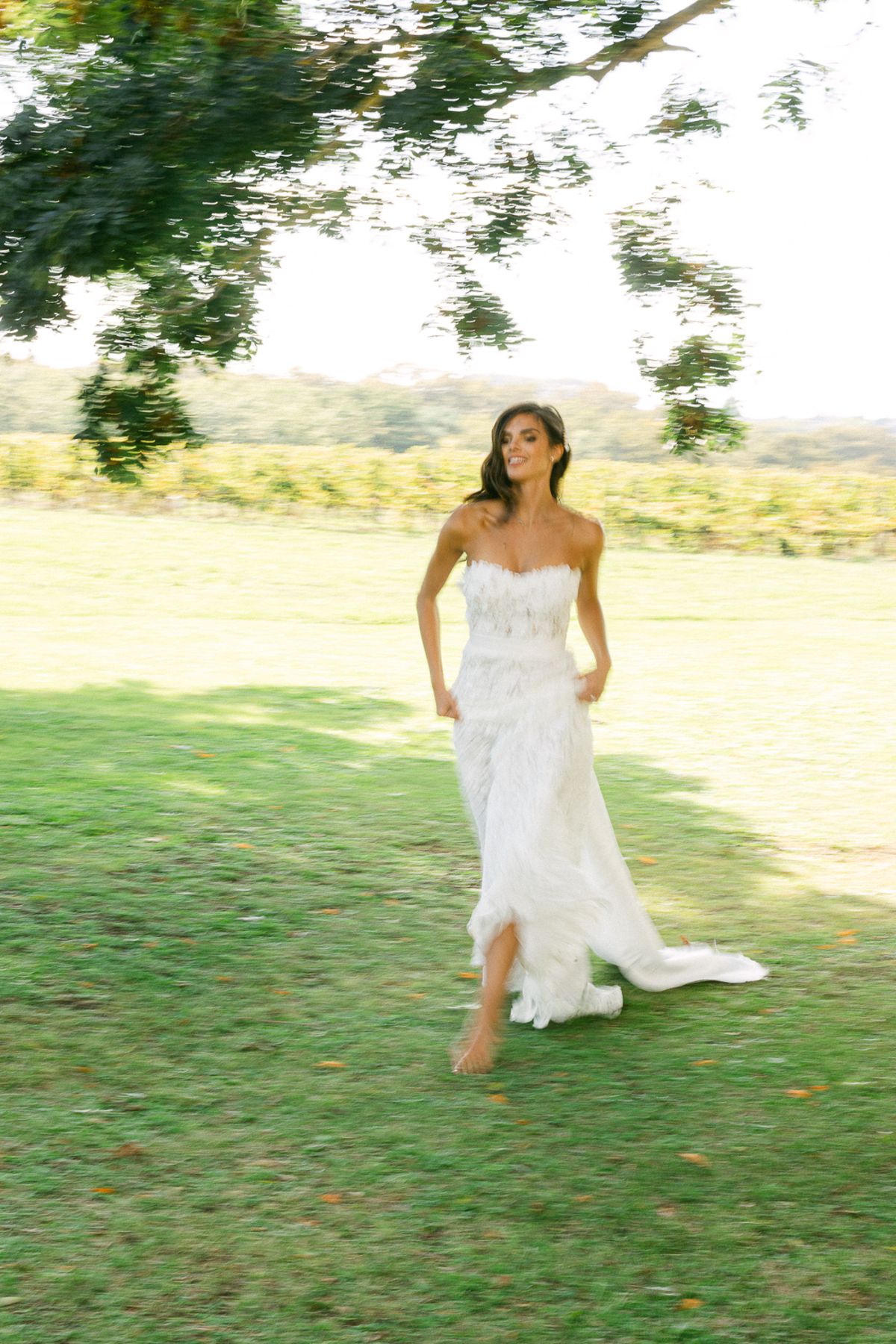 The bride in a strapless lace gown walks barefoot across a sunlit lawn, with vines and trees behind her.