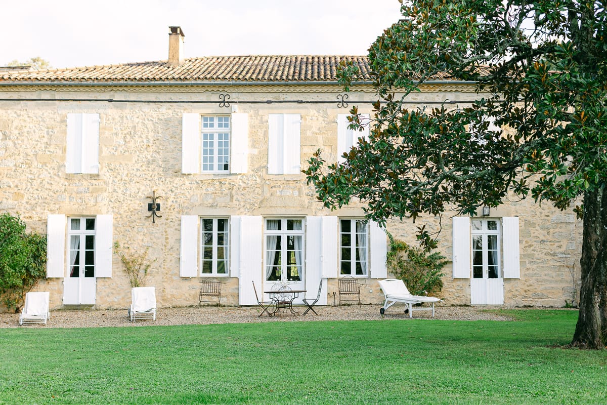 A honey-coloured stone house with white shuttered windows and French doors faces a lawn, with a large tree to the right and chairs by the wall.