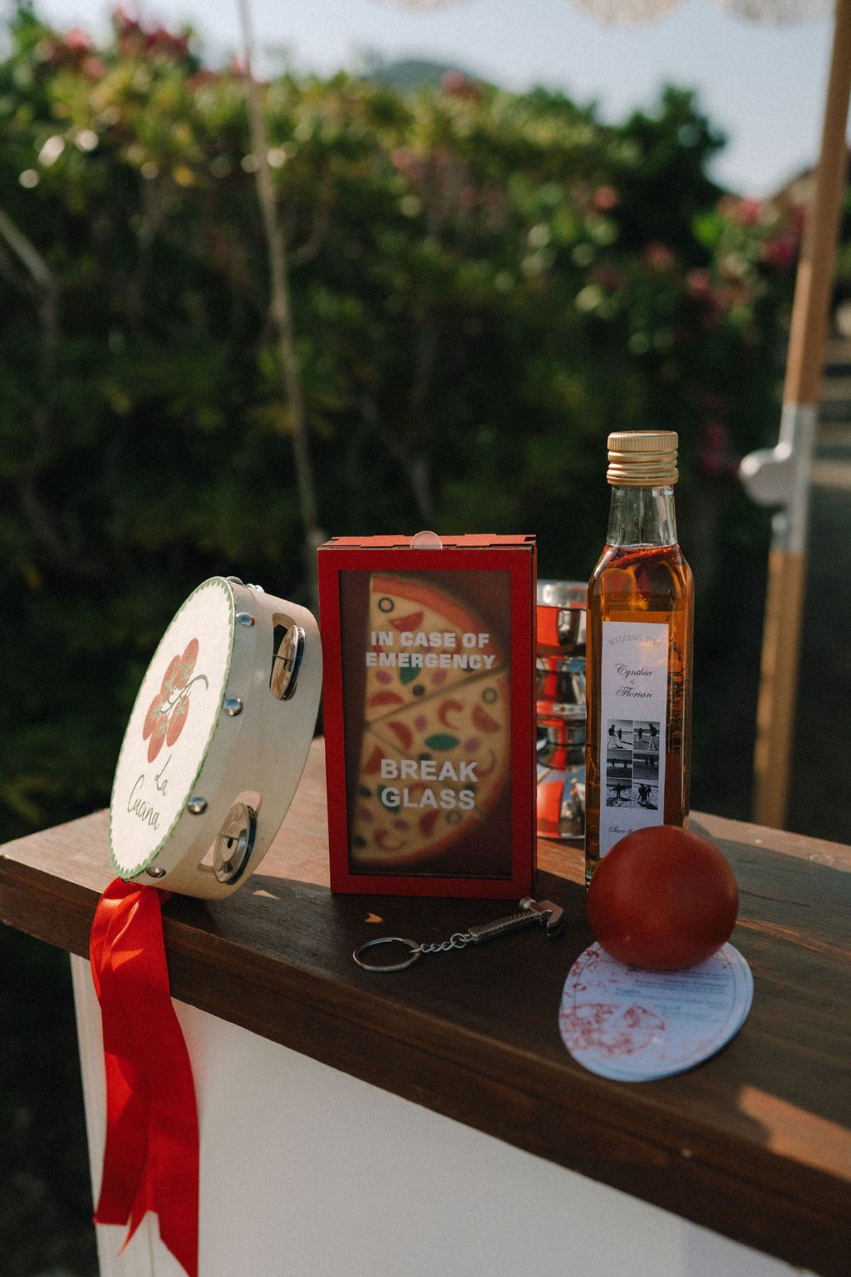 A wooden counter holds a tambourine with a red ribbon, a red box marked break glass, a bottle of oil and a tomato.