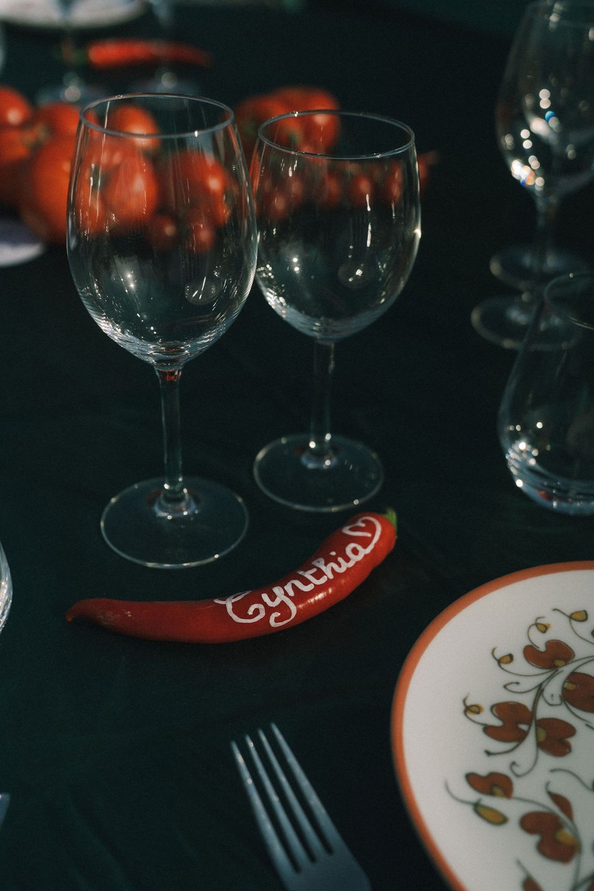 A dark table is set with wine glasses, a patterned plate, small red tomatoes and a name card shaped like a red chilli.