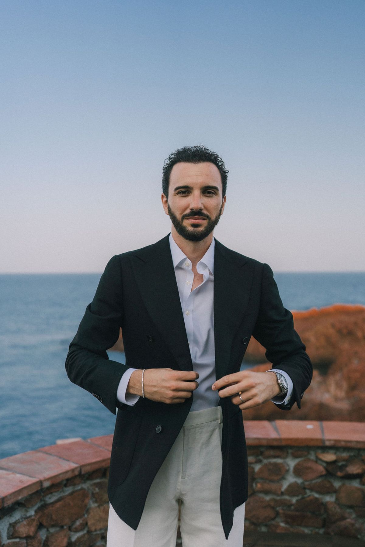 The groom in a navy jacket and light trousers stands adjusting his lapel on a terrace, with the sea behind him.