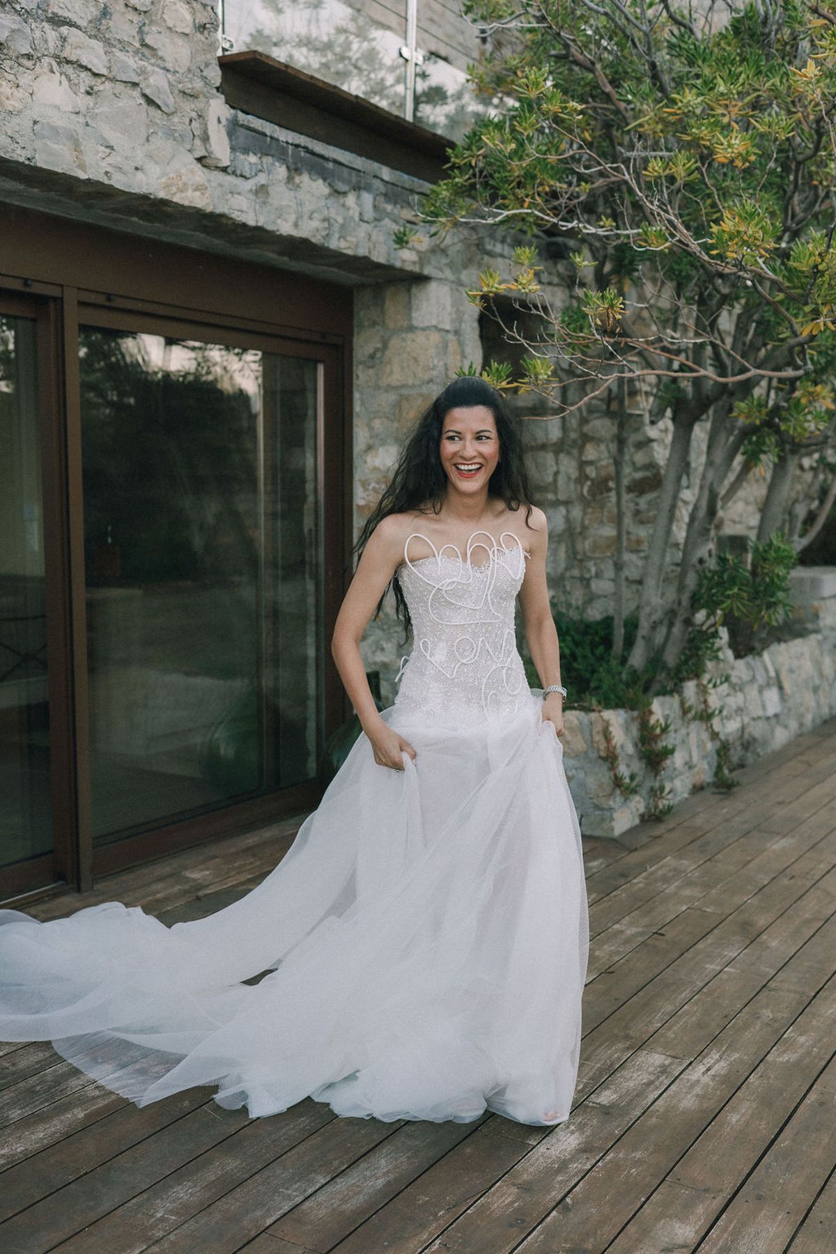 The bride smiles on a wooden deck in front of a stone building, her long train spread behind her.