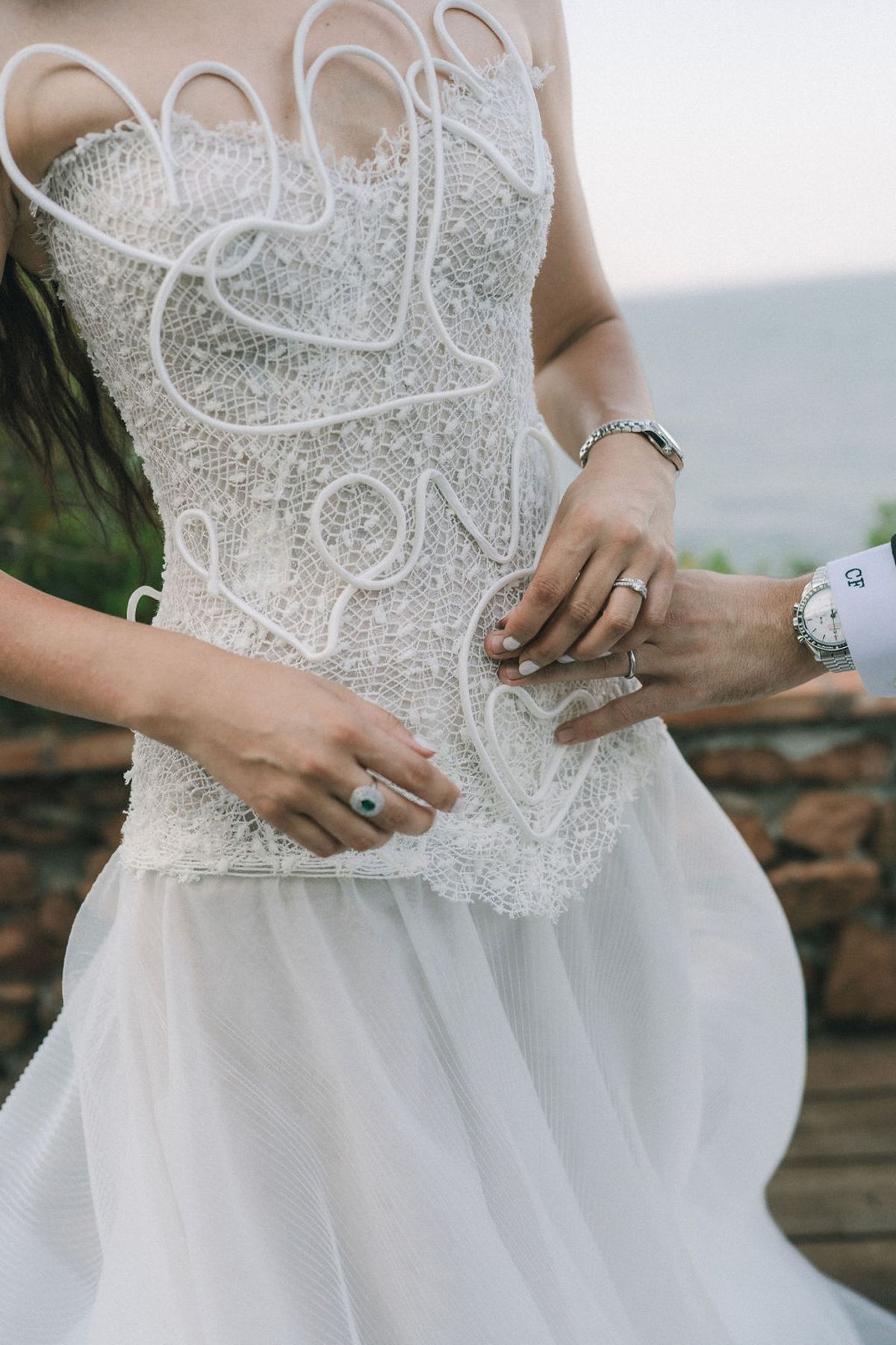 The bride's embroidered bodice and tulle skirt, with her hands and the groom's wrist, wearing rings and a watch, by the sea.