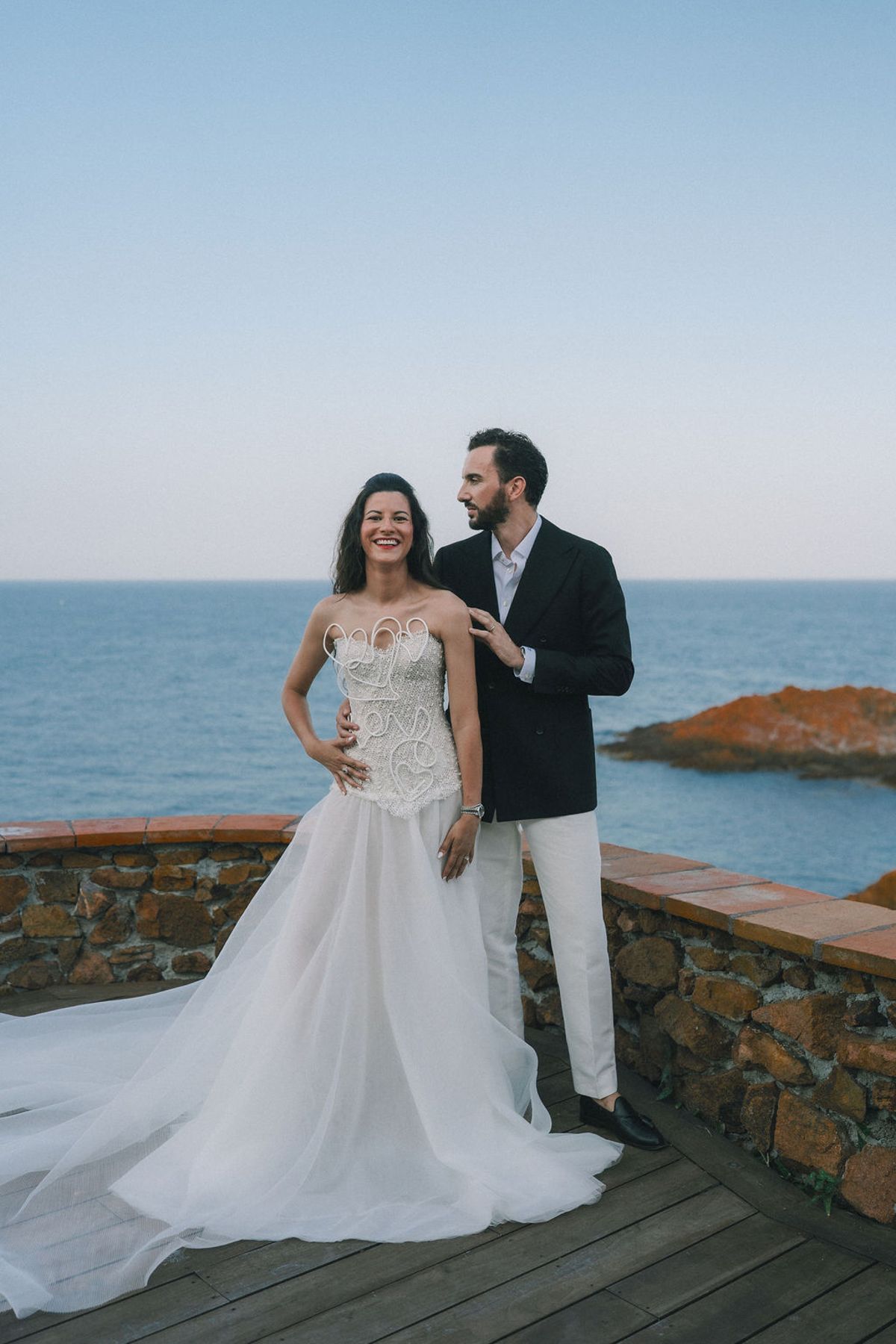 The groom in a dark jacket stands close to the bride in her gown, with the sea and a clear sky behind them.
