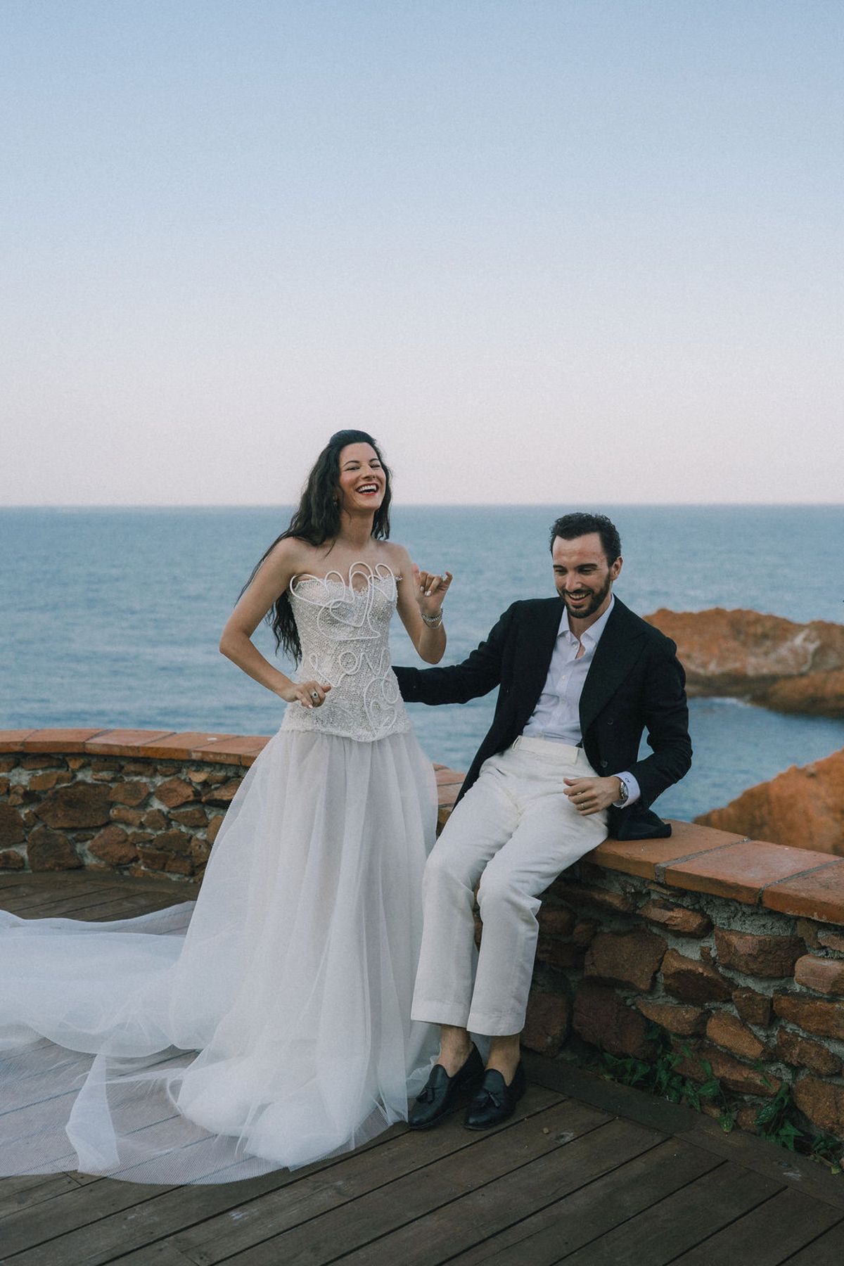 The bride in a strapless gown stands laughing beside the groom, who sits on a stone wall with the sea behind them.