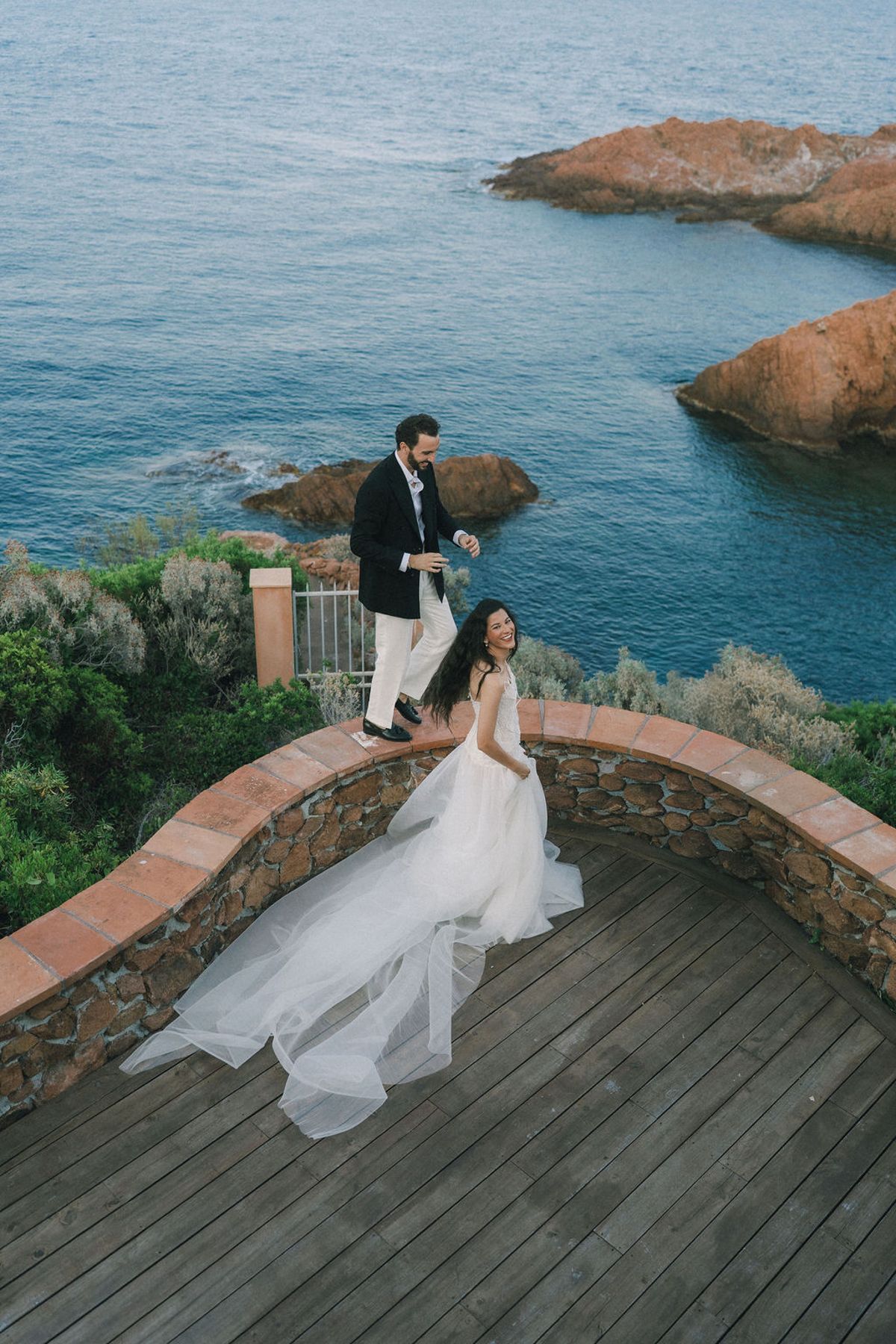 The groom stands on a low stone wall above the sea while the bride sits below, her long tulle train spread across the deck.