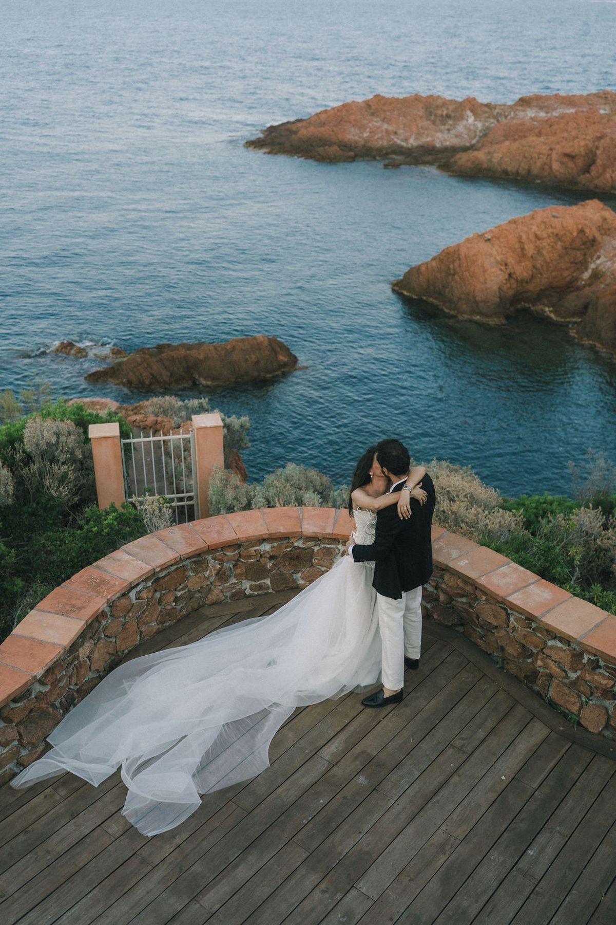 Seen from above, the couple embrace on a wooden terrace, the bride's long veil trailing, above a rocky blue coastline.