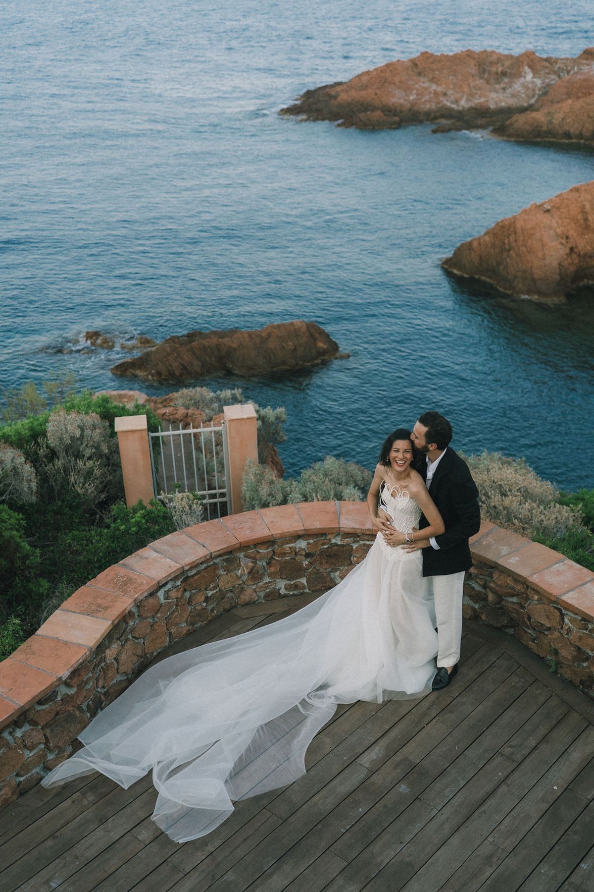 Seen from above, the couple embrace on a wooden terrace, the bride's long train trailing, with rocks and blue sea behind.