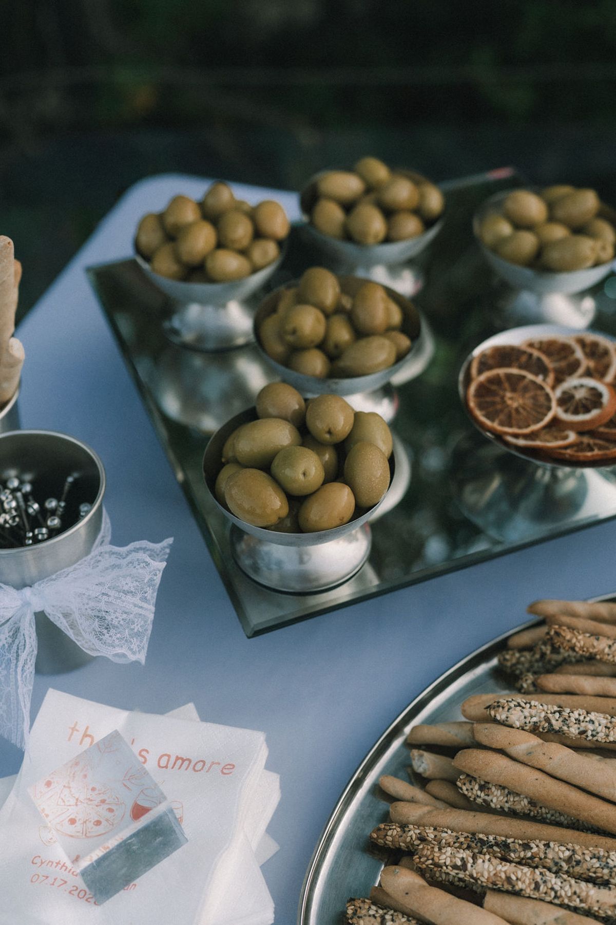Small bowls of green olives, dried orange slices and a tray of breadsticks sit on a table with a lace-trimmed napkin.