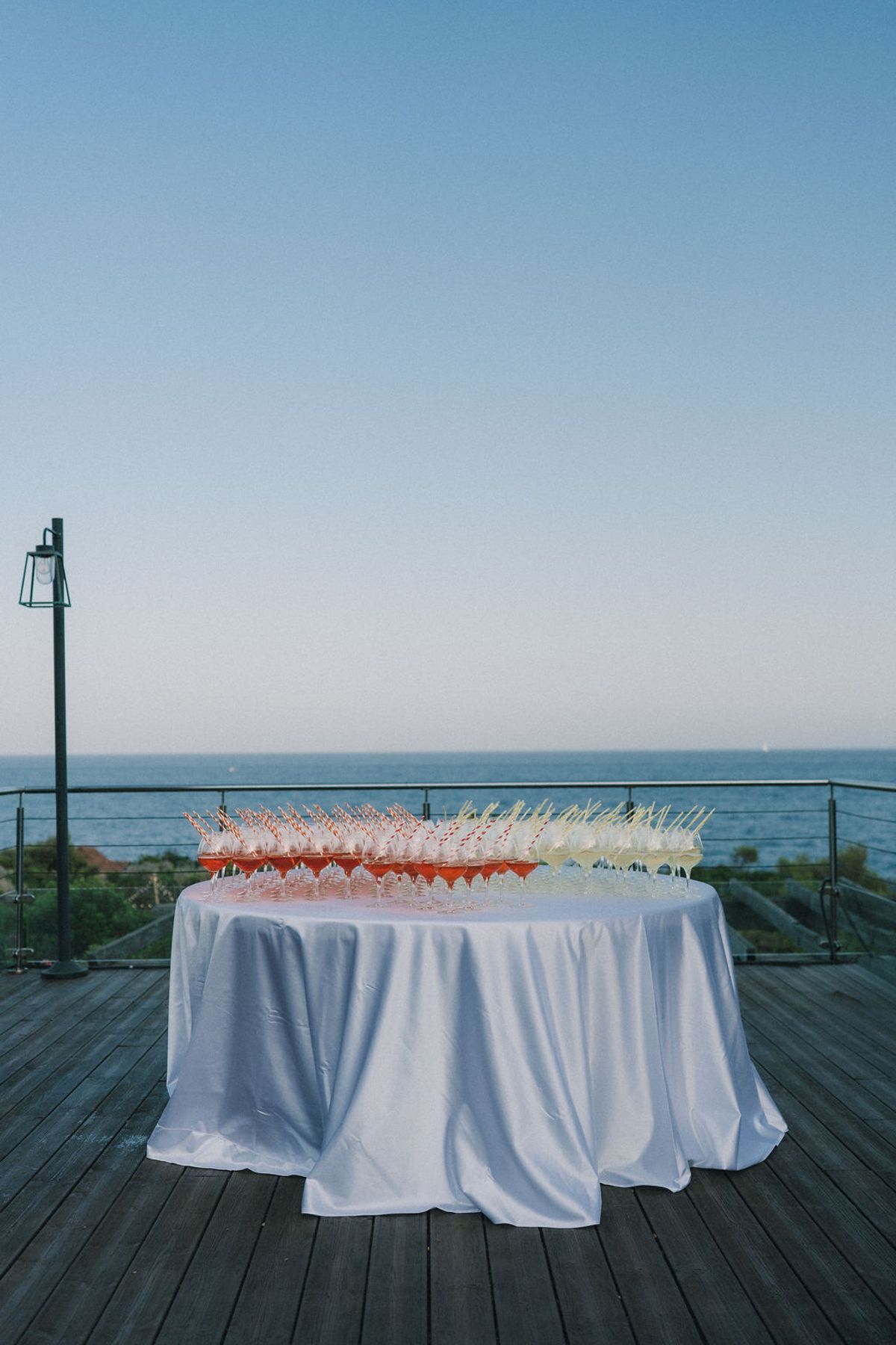 A round table with a white cloth holds many filled champagne glasses on a wooden deck, with the sea and a clear sky behind.