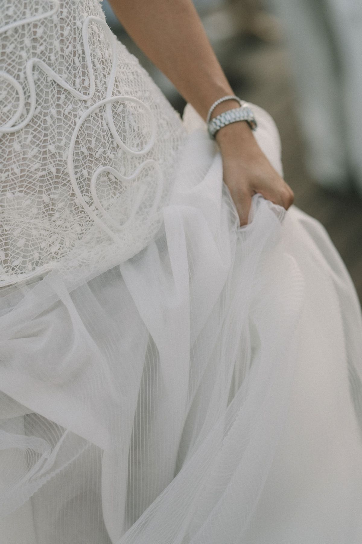 The bride holds the layered tulle skirt of her gown, with a beaded bodice and a bracelet visible, her face out of frame.