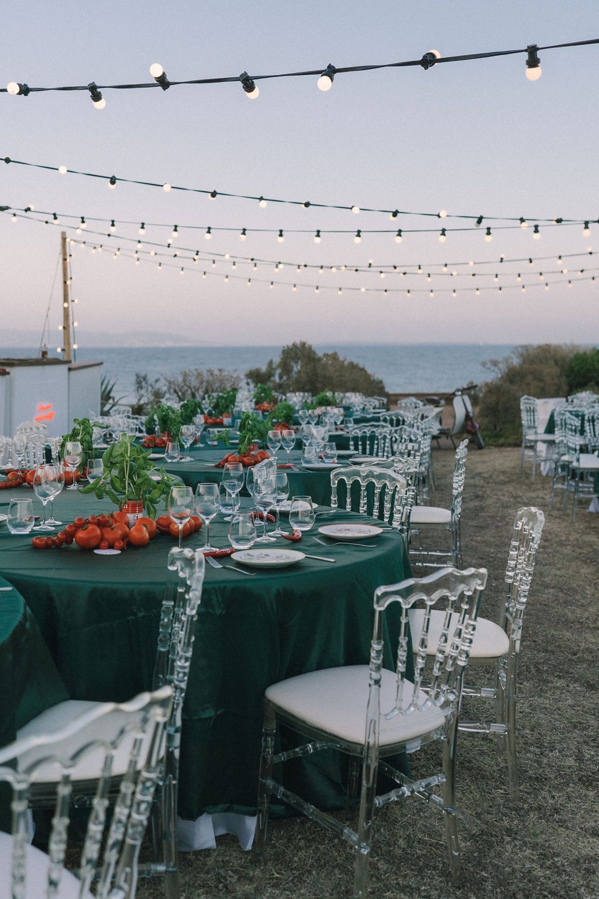 Round tables with dark green cloths and clear chairs are laid out beneath strings of lights, with the sea beyond.