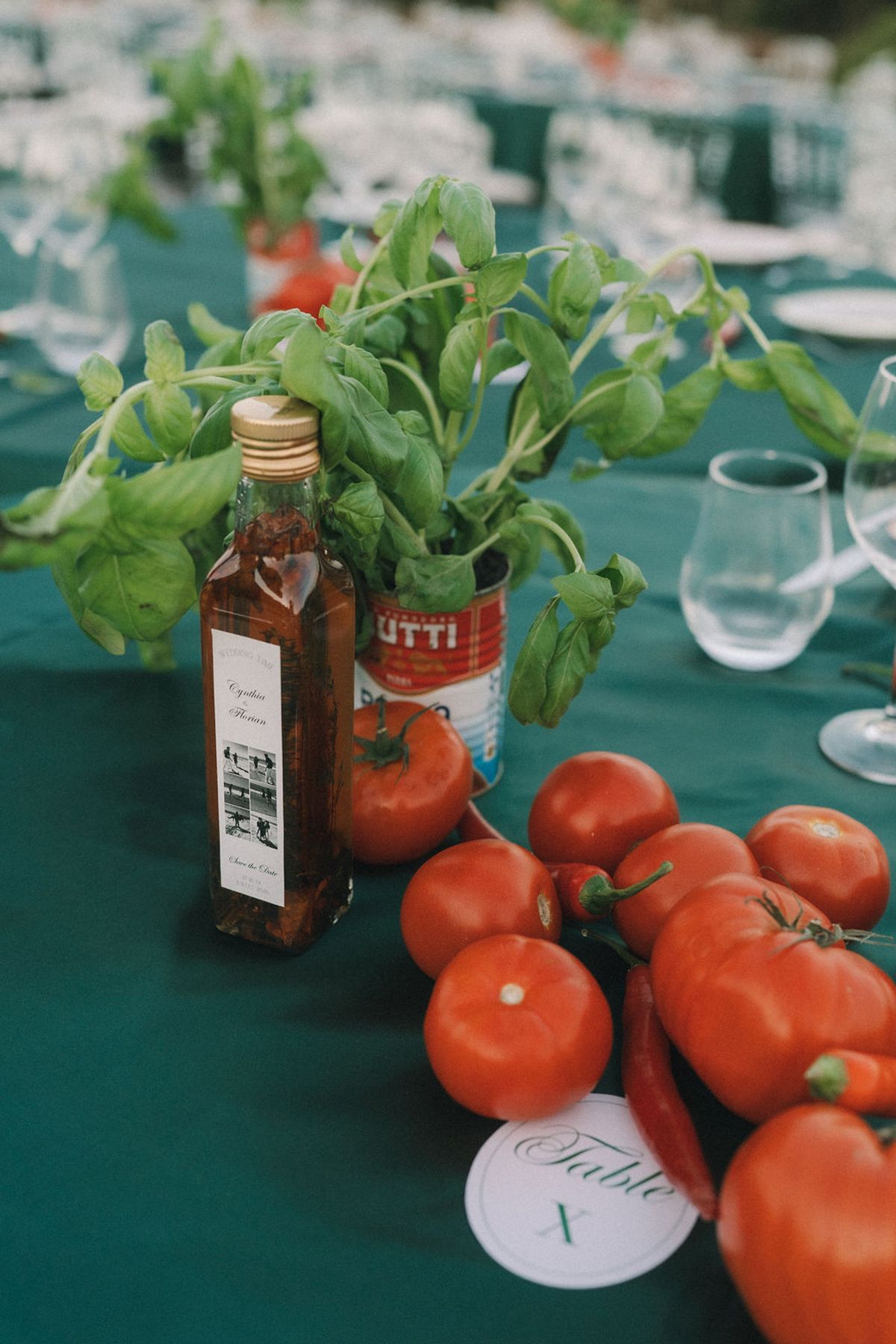 On a green tablecloth, basil grows from a tomato tin beside an oil bottle, fresh tomatoes, a glass and a table number card.