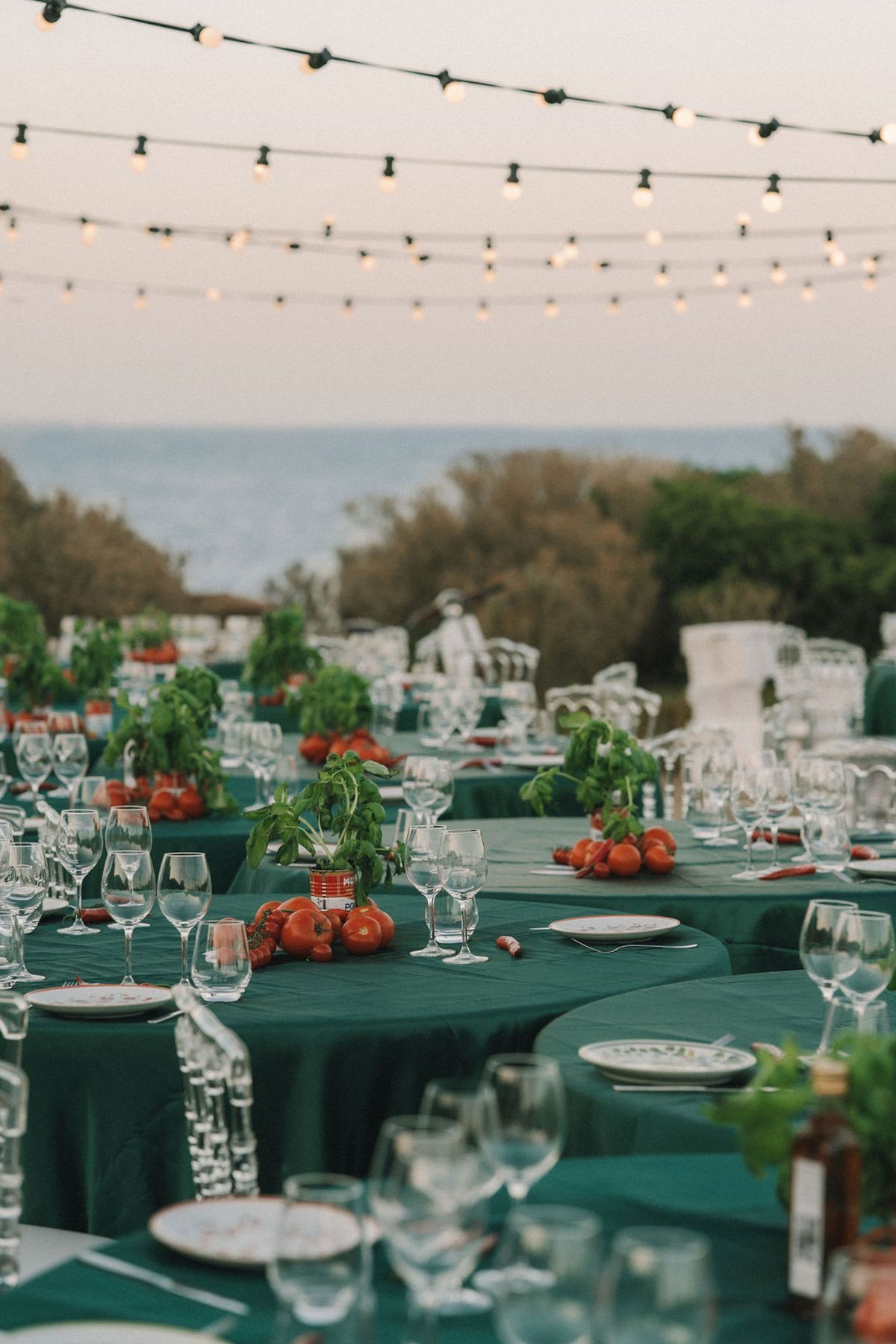 Tables with green cloths hold glasses, tomatoes and herb plants, with hanging lights and the sea in the distance.