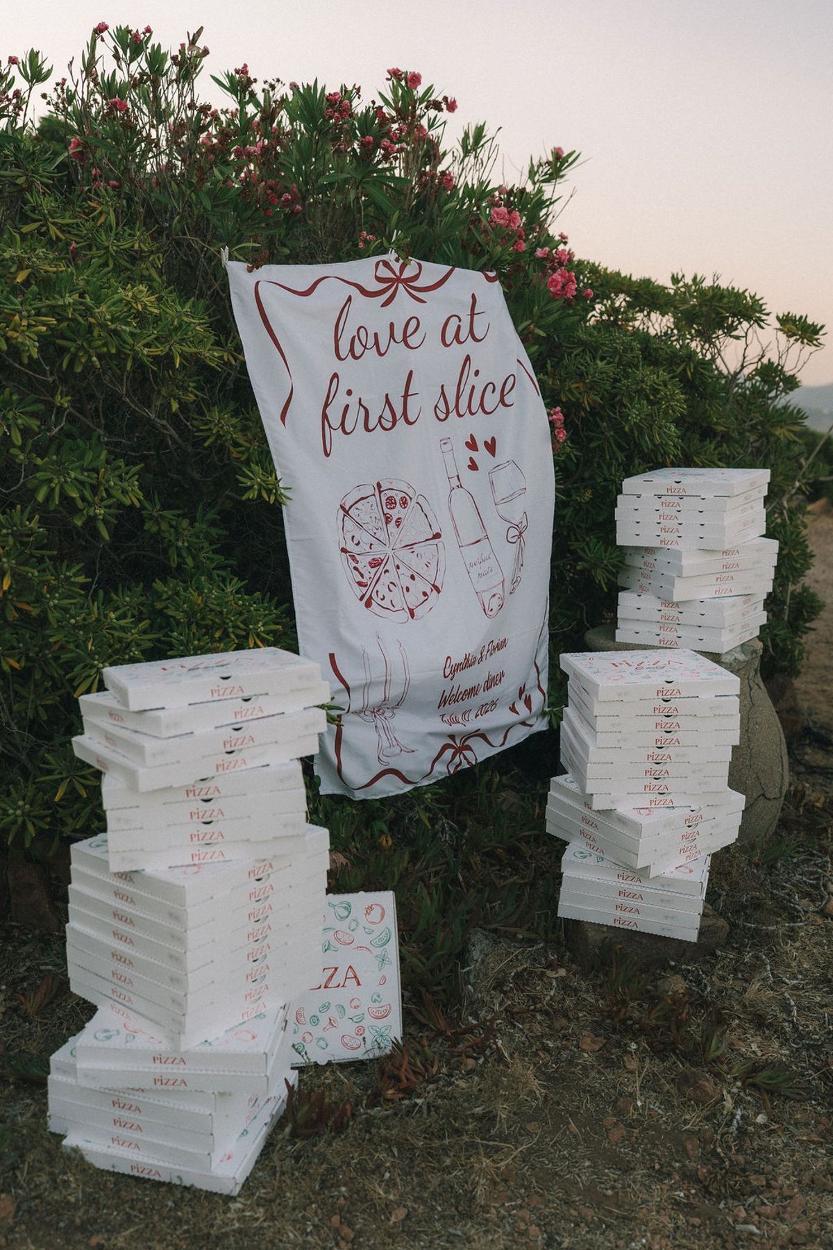 A printed banner reading love at first slice stands beside stacks of white pizza boxes among coastal shrubs.