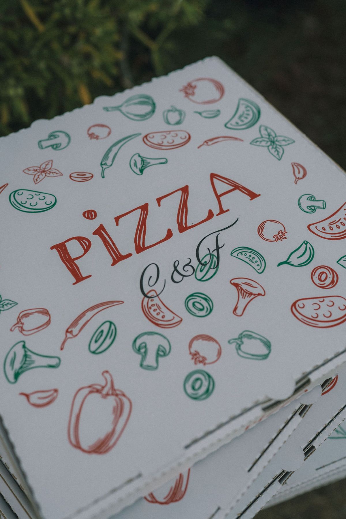 A stack of white pizza boxes printed with colourful vegetable illustrations, the word pizza in red and a pair of initials.