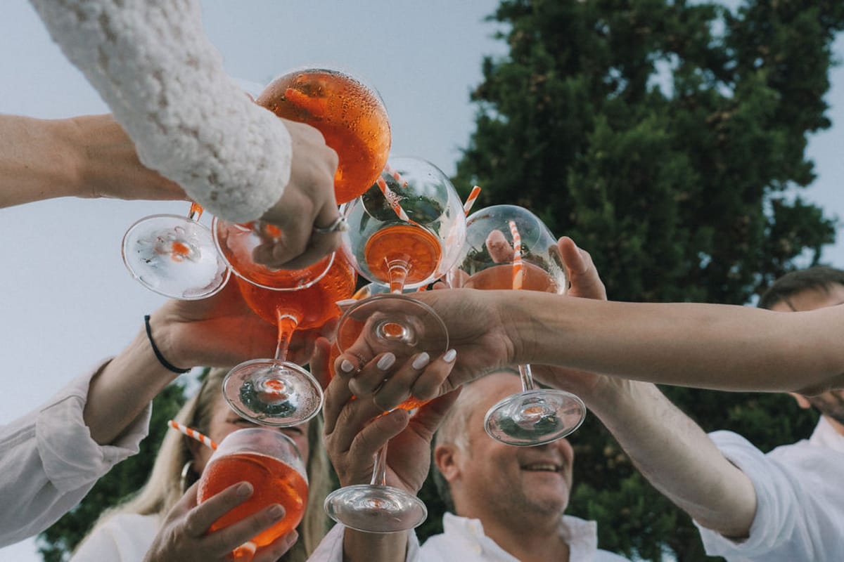 Several guests' hands raise orange aperitif glasses together in a toast beneath a blue sky and trees.
