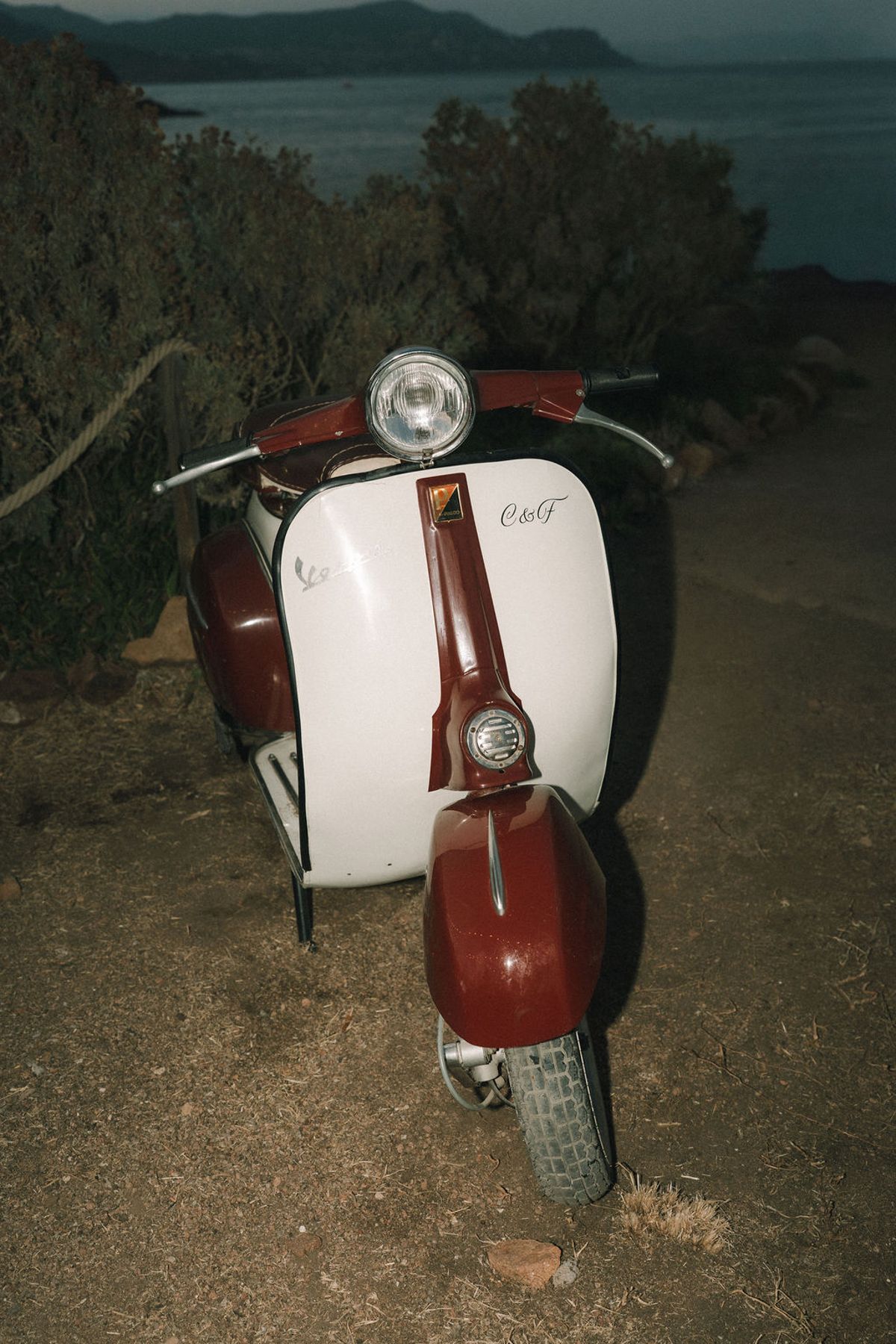 A vintage red and white scooter is parked on dry ground among bushes, with the sea visible in the dim distance.
