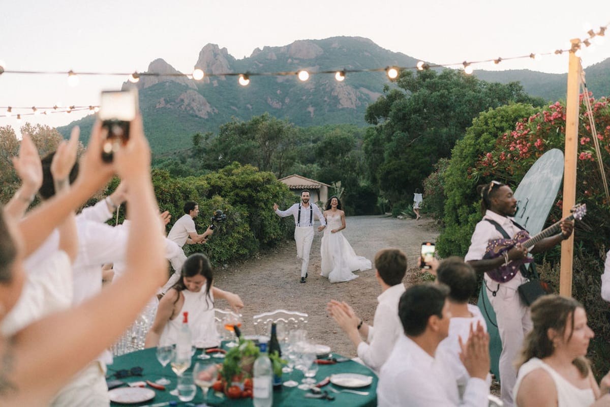The couple walks along a gravel path toward guests dressed in white, who clap and film, with string lights and hills behind.