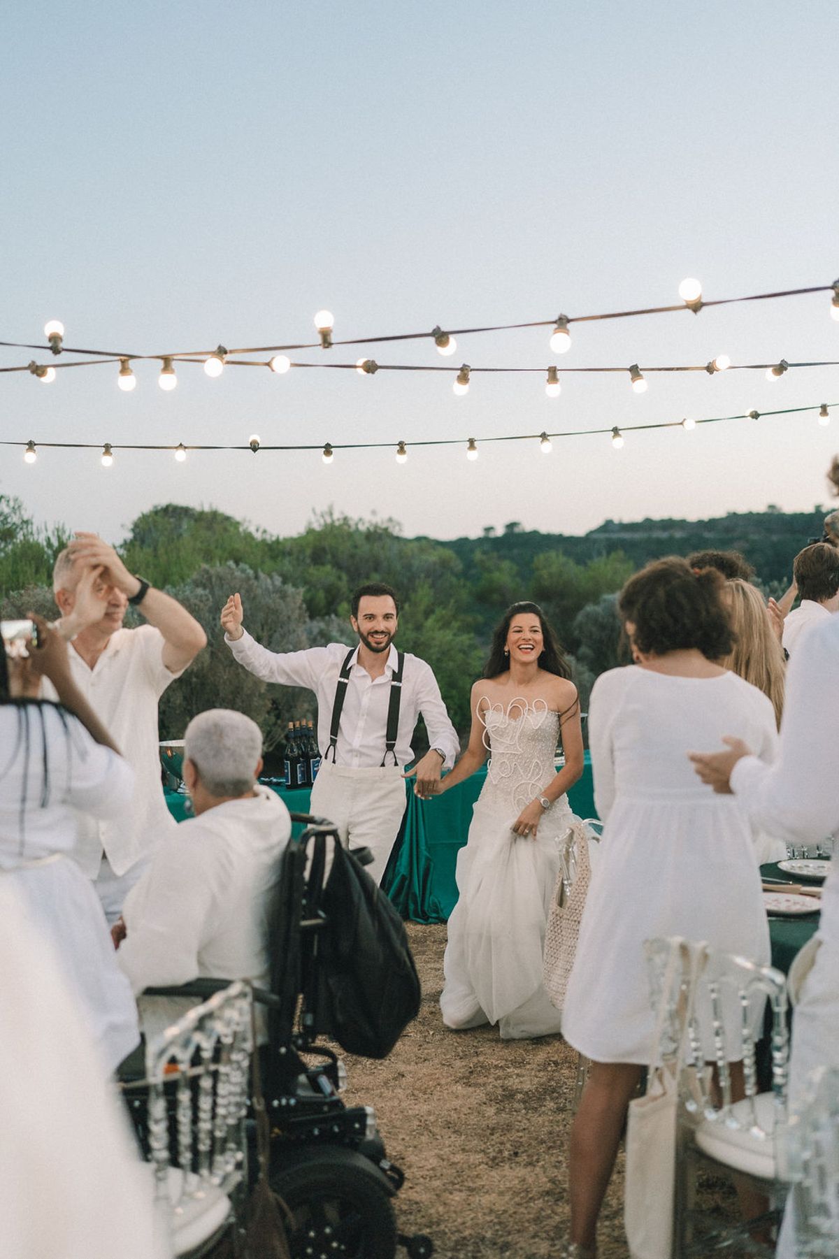 The groom and bride walk hand in hand among guests in white who dance under string lights at dusk.