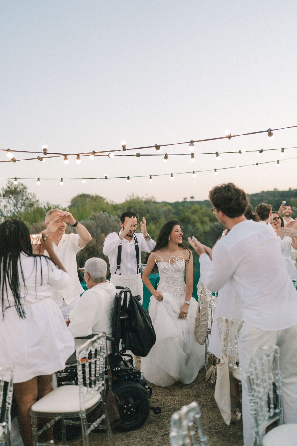Guests dressed in white raise their arms and cheer around the bride beneath strings of lights, with one guest seated in a wheelchair.