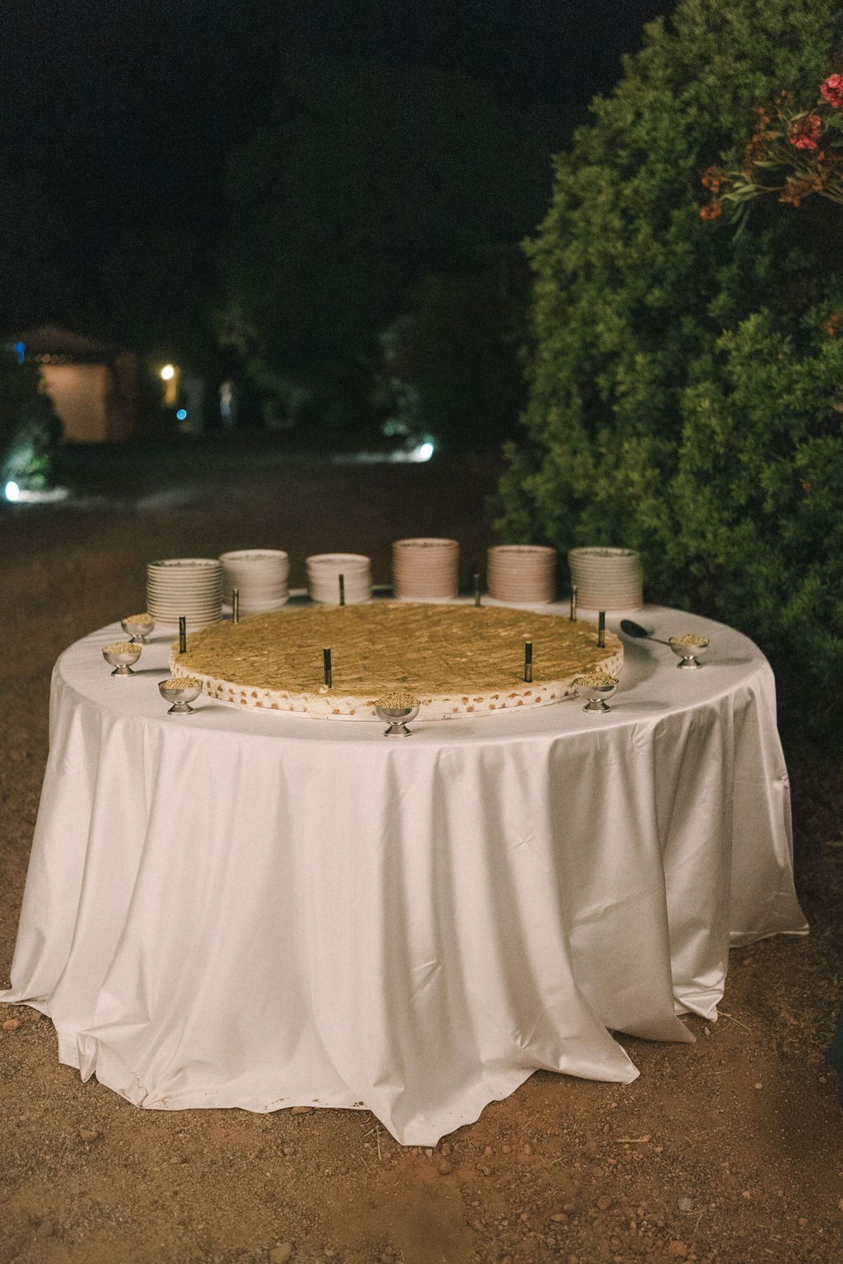 A round table with a white cloth holds a golden tart and a ring of small cups, set outdoors at night.