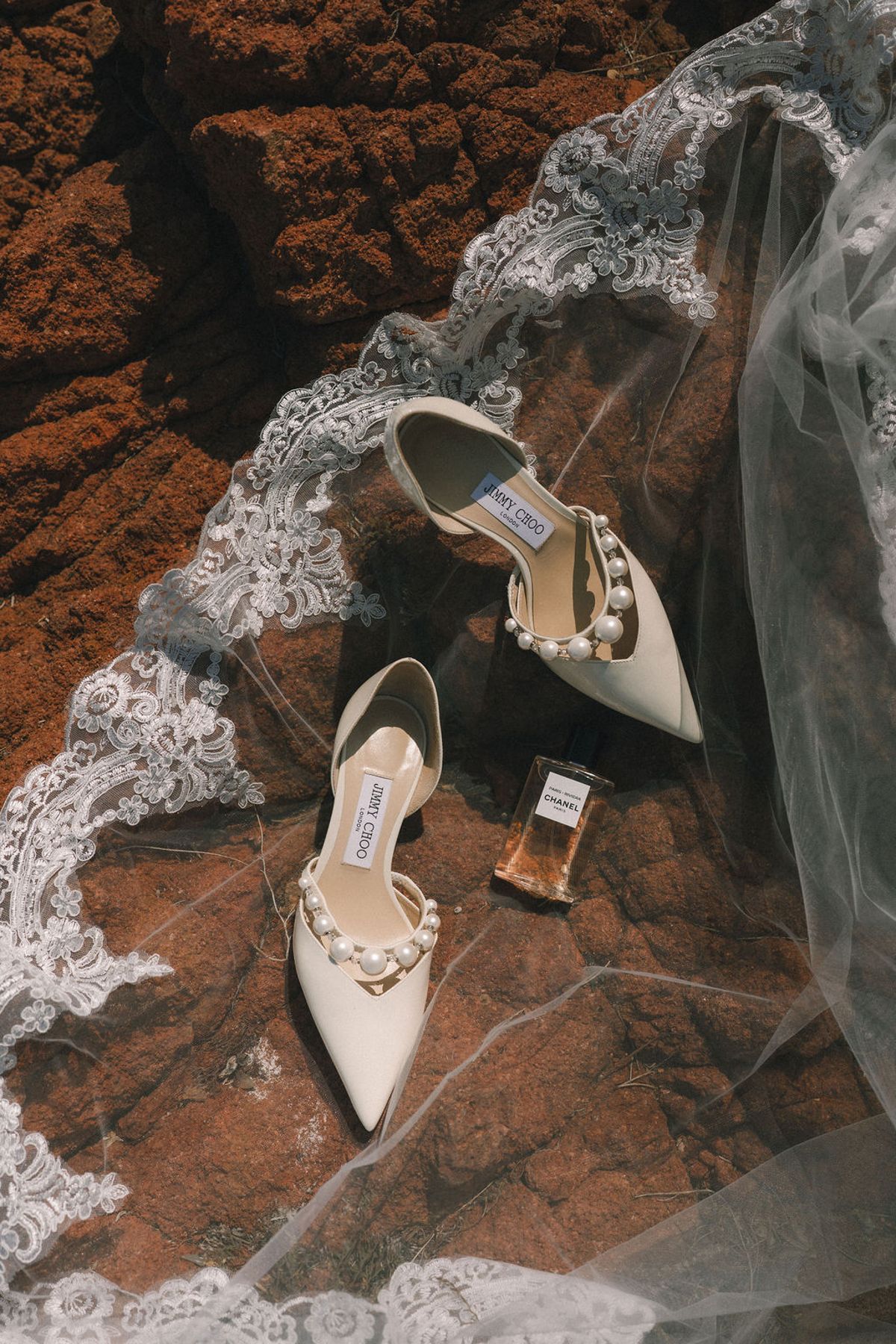 A pair of pearl-trimmed bridal shoes and a small perfume bottle rest on red rock, partly covered by a lace-edged veil.