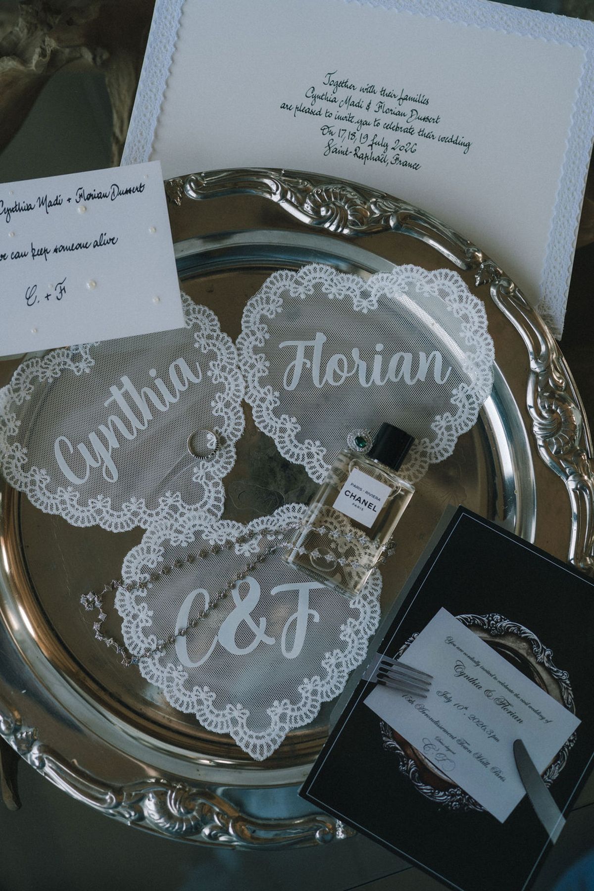 Overhead view of a silver tray holding lace heart-shaped name cards, a perfume bottle, an invitation and a black card.