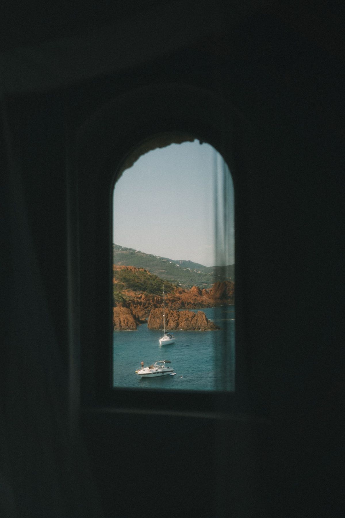 From a dark stone room, an arched window frames a bay with red rocks, blue water and small boats.