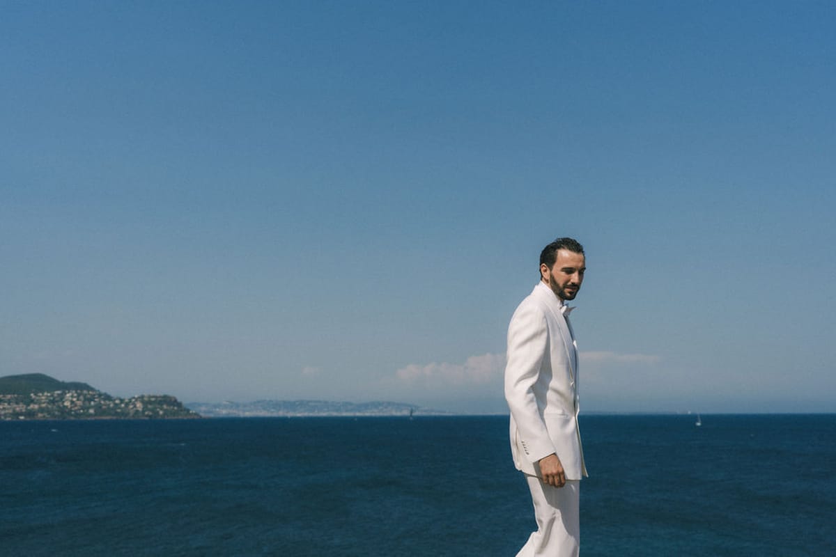 The groom in a white suit stands side-on against a deep blue sea and clear sky, with a distant coastline.