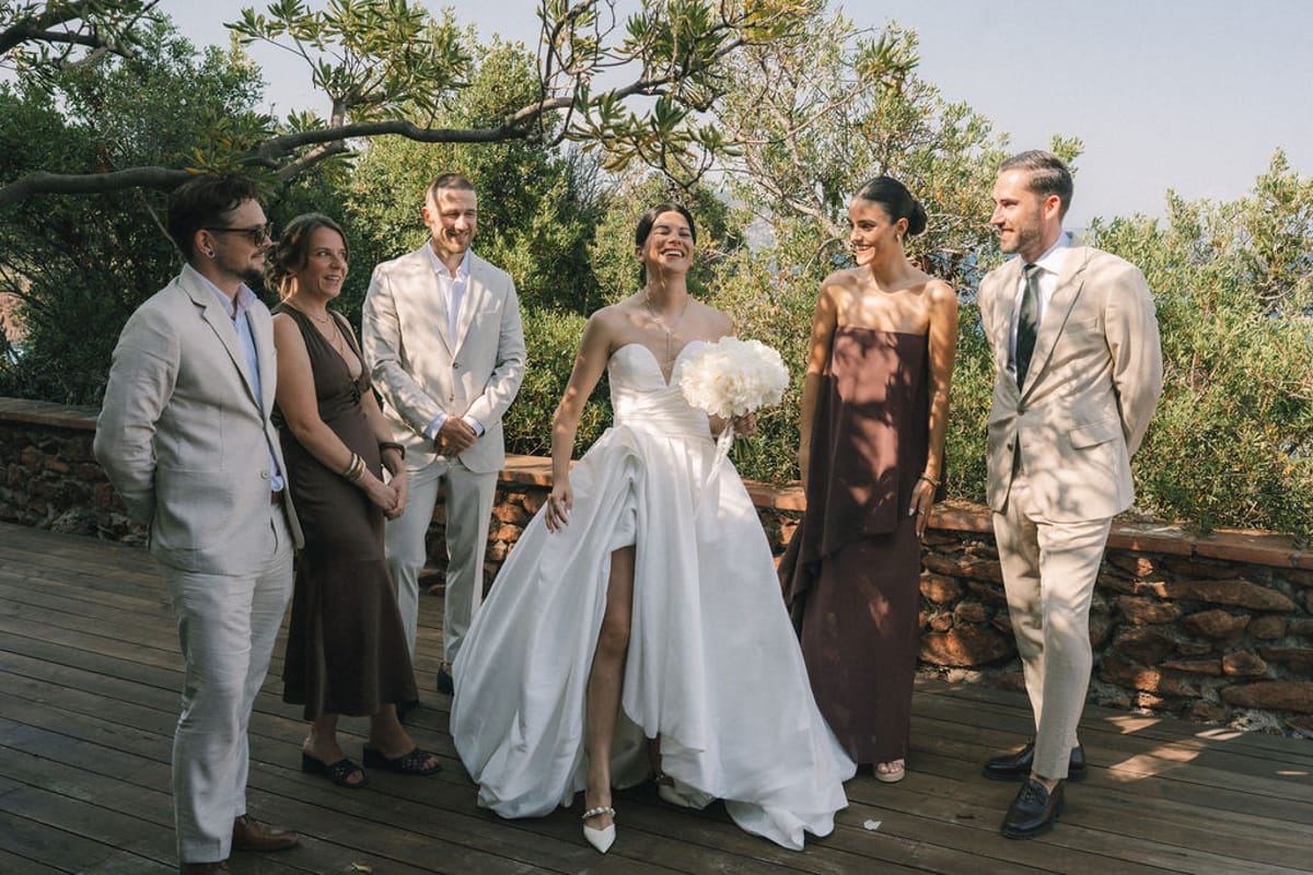 The bride, in a white gown with a bouquet, stands laughing with five companions in neutral and brown outfits on a wooden terrace.