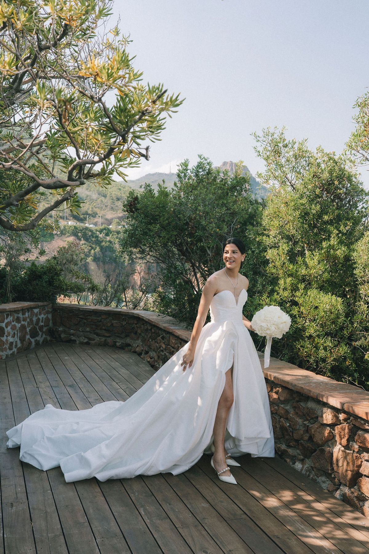 The bride stands on a wooden terrace holding a white bouquet, her long gown trailing, with green trees and blue sky behind.