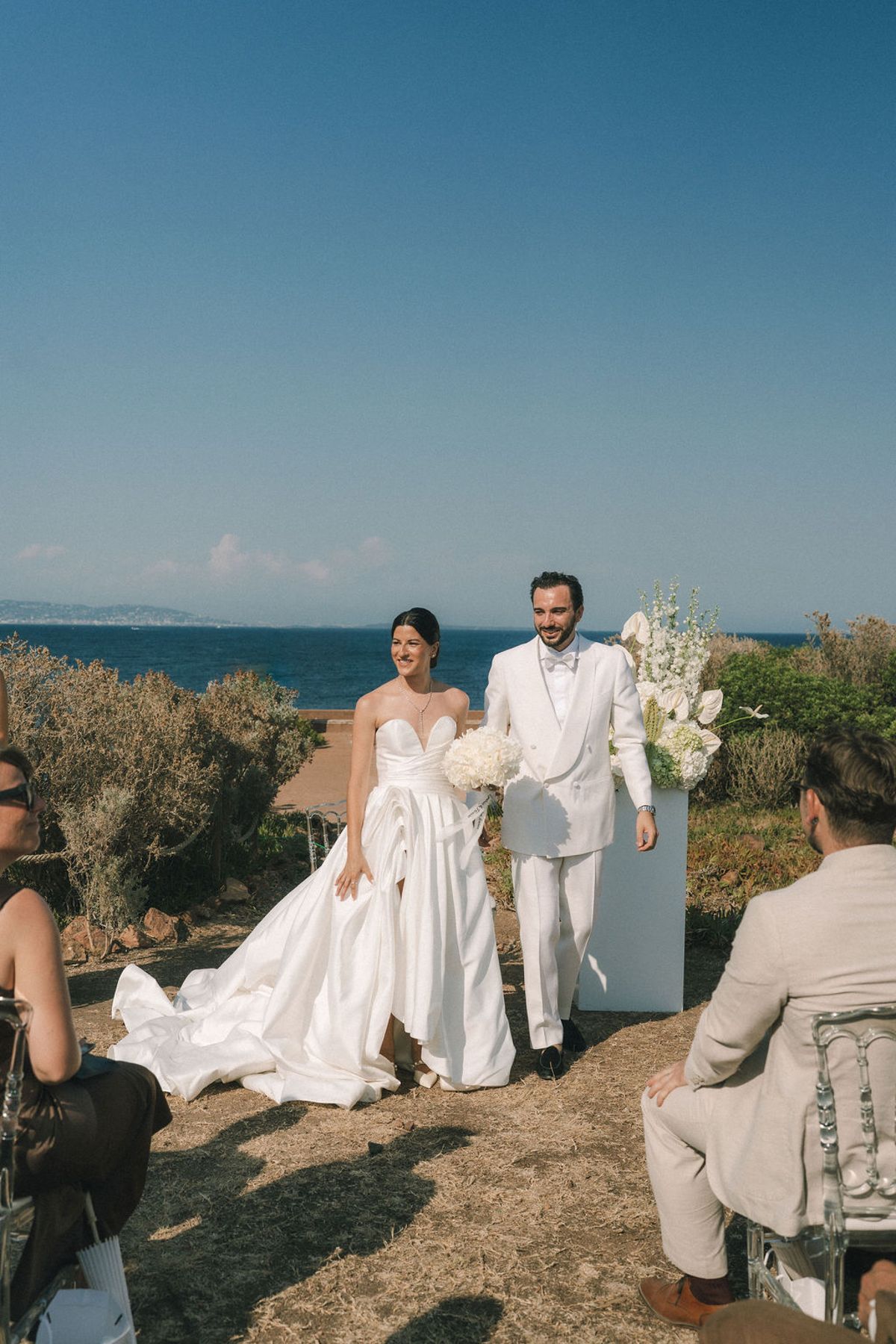 The bride in a white gown with a long train and the groom in a white suit walk past seated guests by the sea.